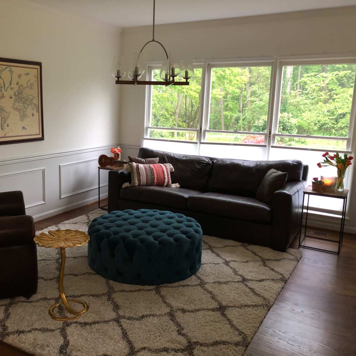 A cozy living room features a brown leather sofa, a blue tufted ottoman, and a vintage world map on the wall, with large windows showcasing a view of greenery outside.