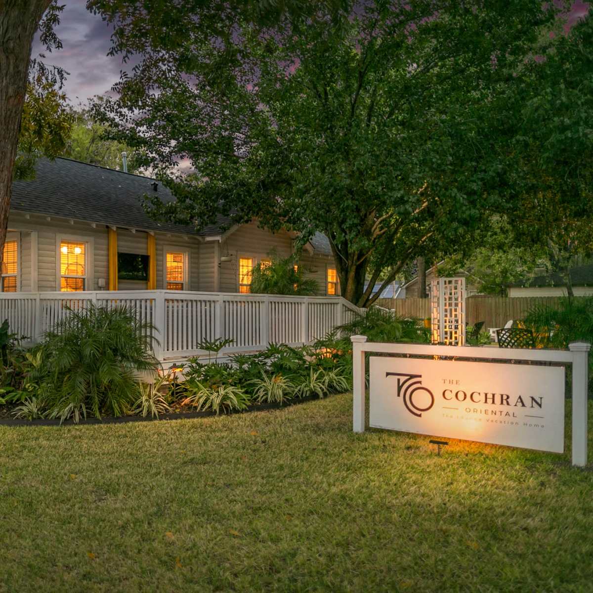 A white house with a porch is surrounded by lush greenery and illuminated by lights, featuring a sign that reads "The Cochran."