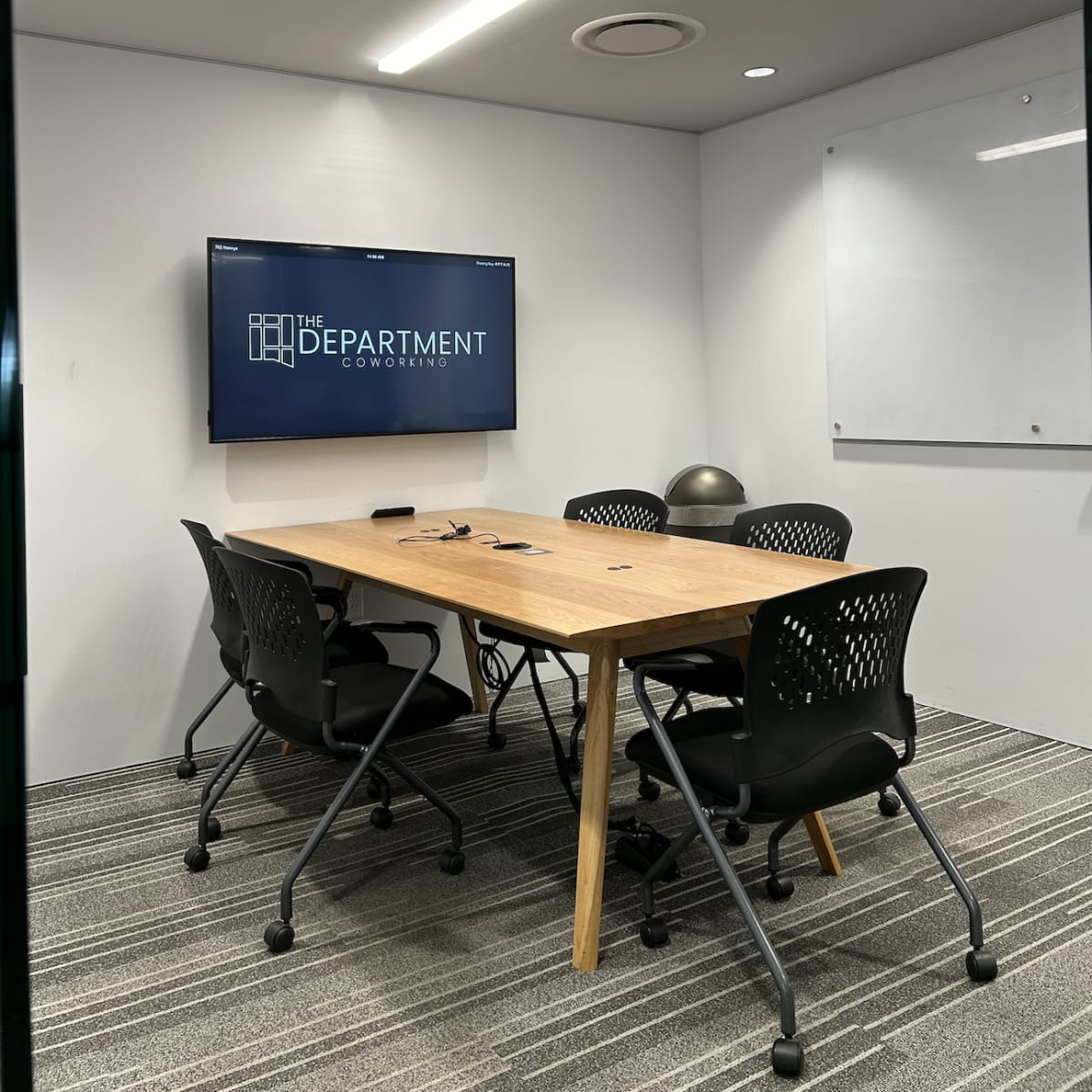 A modern conference room features a wooden table surrounded by four black office chairs, with a large screen displaying "THE DEPARTMENT" on the wall.