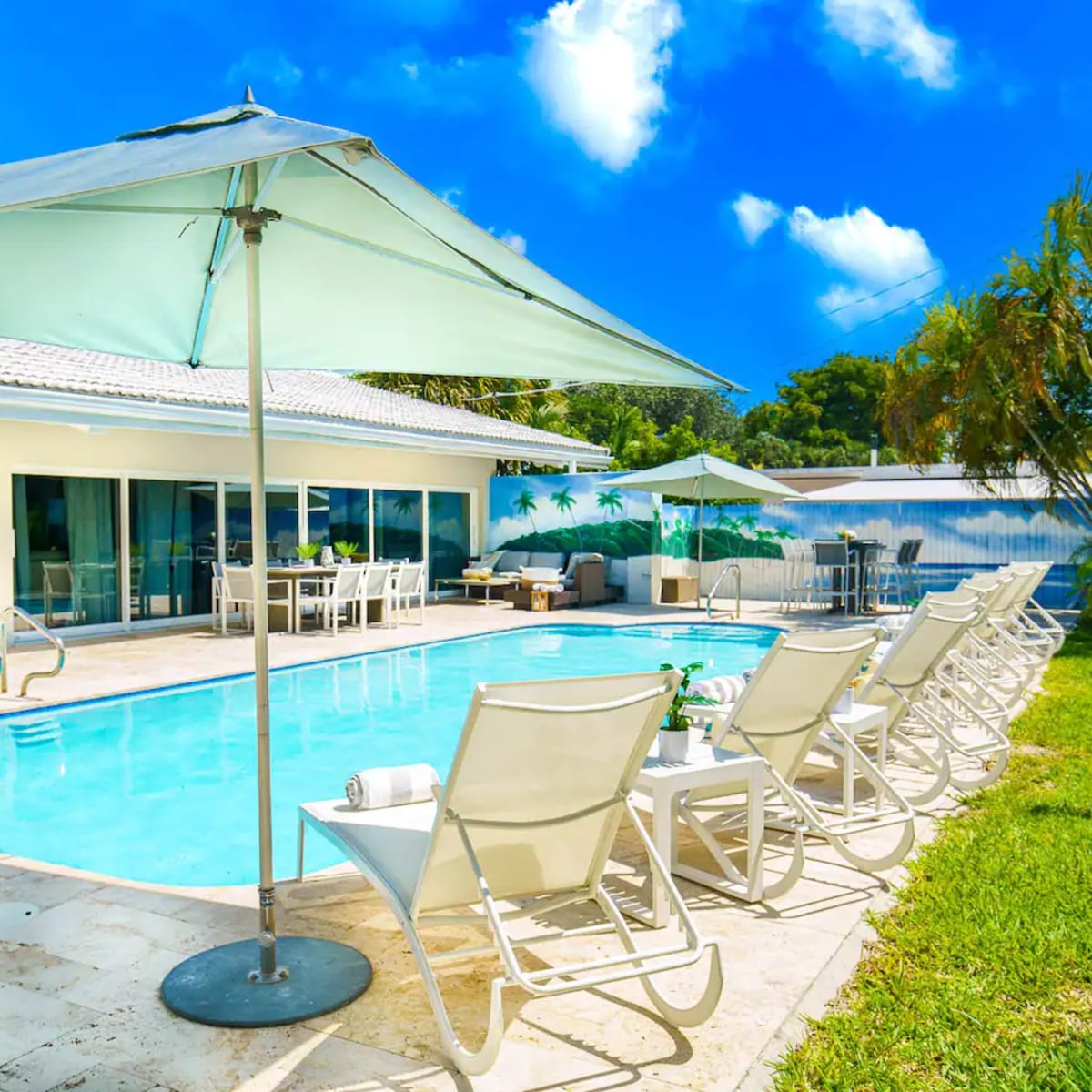 A swimming pool with lounge chairs and umbrellas is surrounded by a lawn and palm trees next to a modern house.
