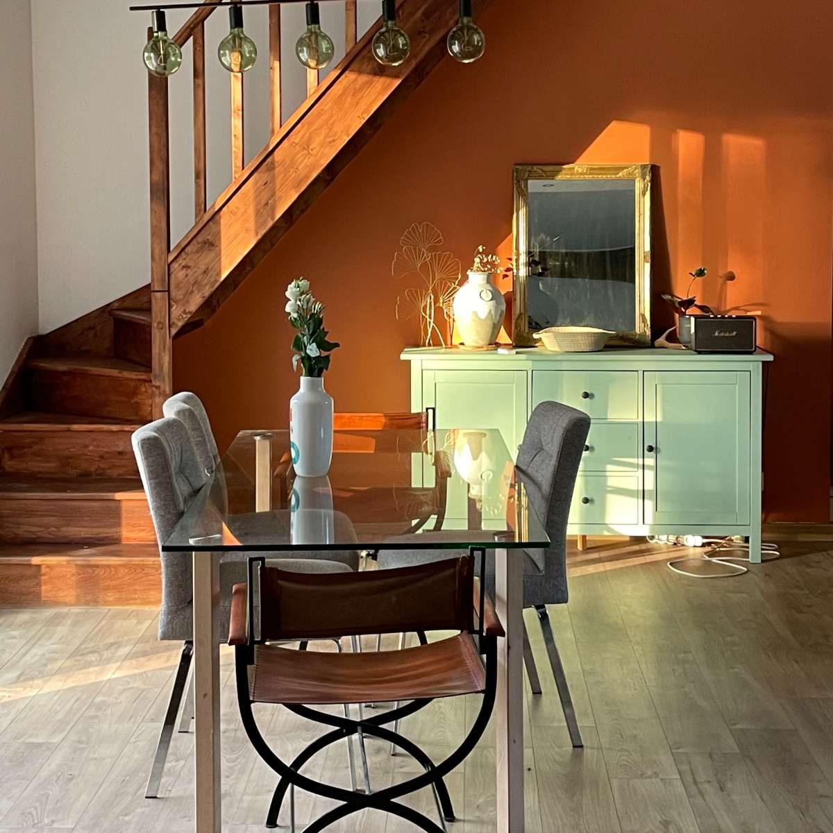 A dining area features a glass table with four chairs, a green sideboard, and a staircase illuminated by pendant lights.
