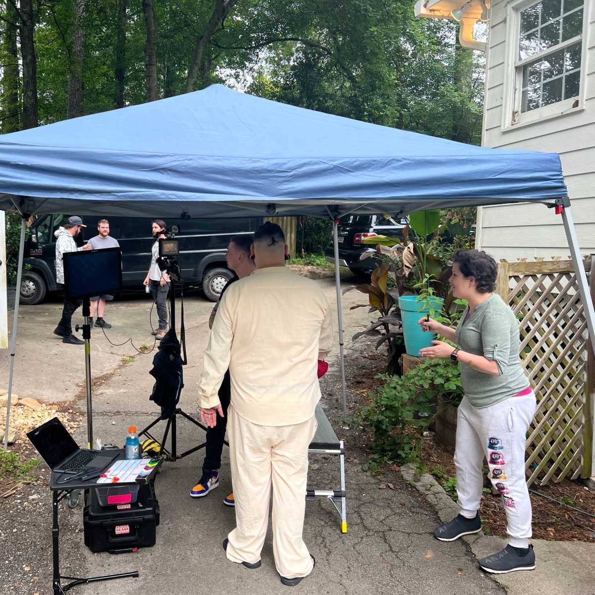 A group of people gathers under a blue tent in a backyard, facing a television setup while a vehicle is parked nearby.