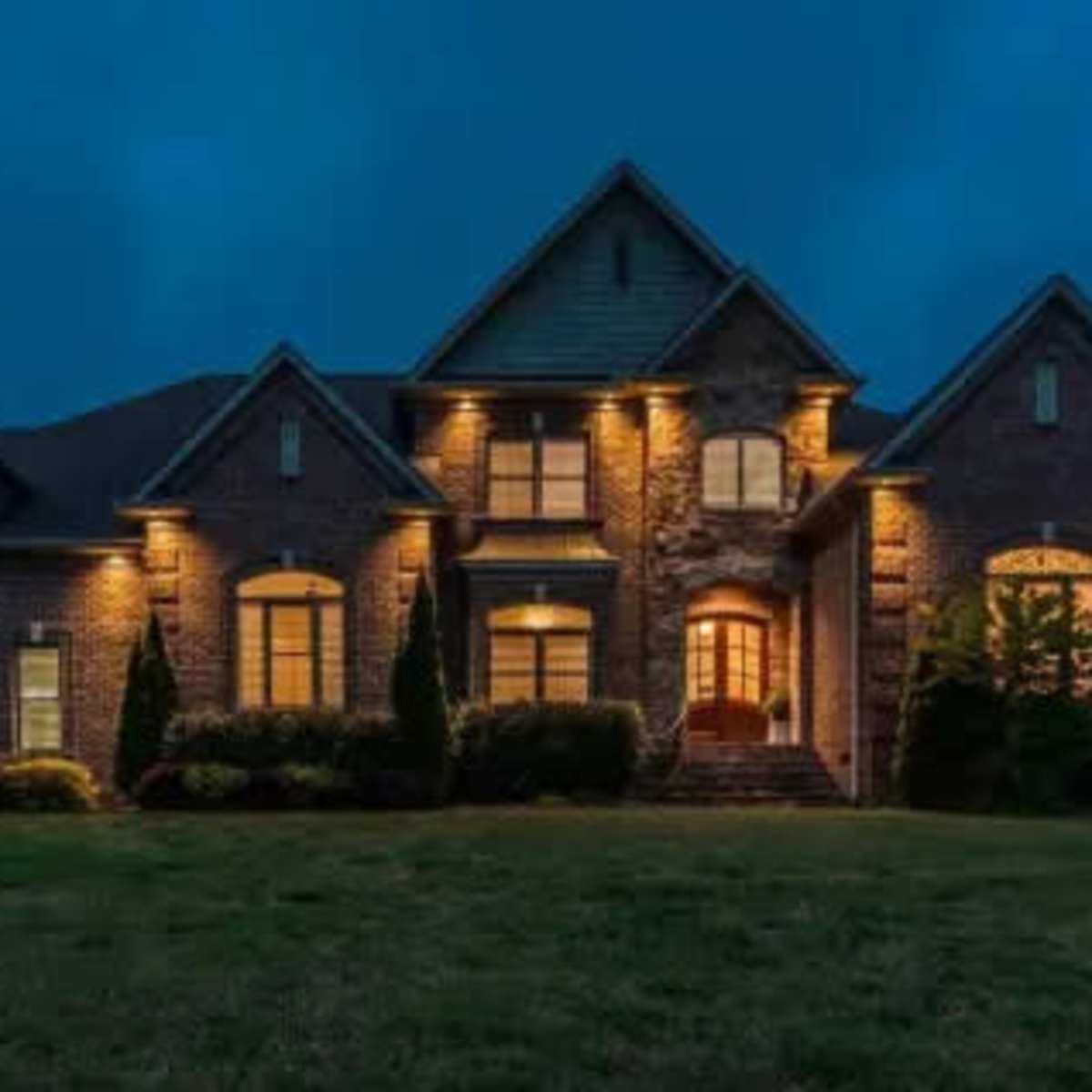 A large brick house illuminated by exterior lights against a twilight sky.