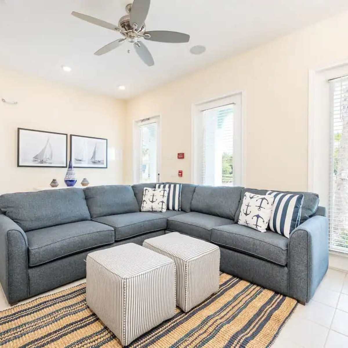 A cozy living room with a gray sectional sofa, two striped ottomans, and windows letting in natural light.
