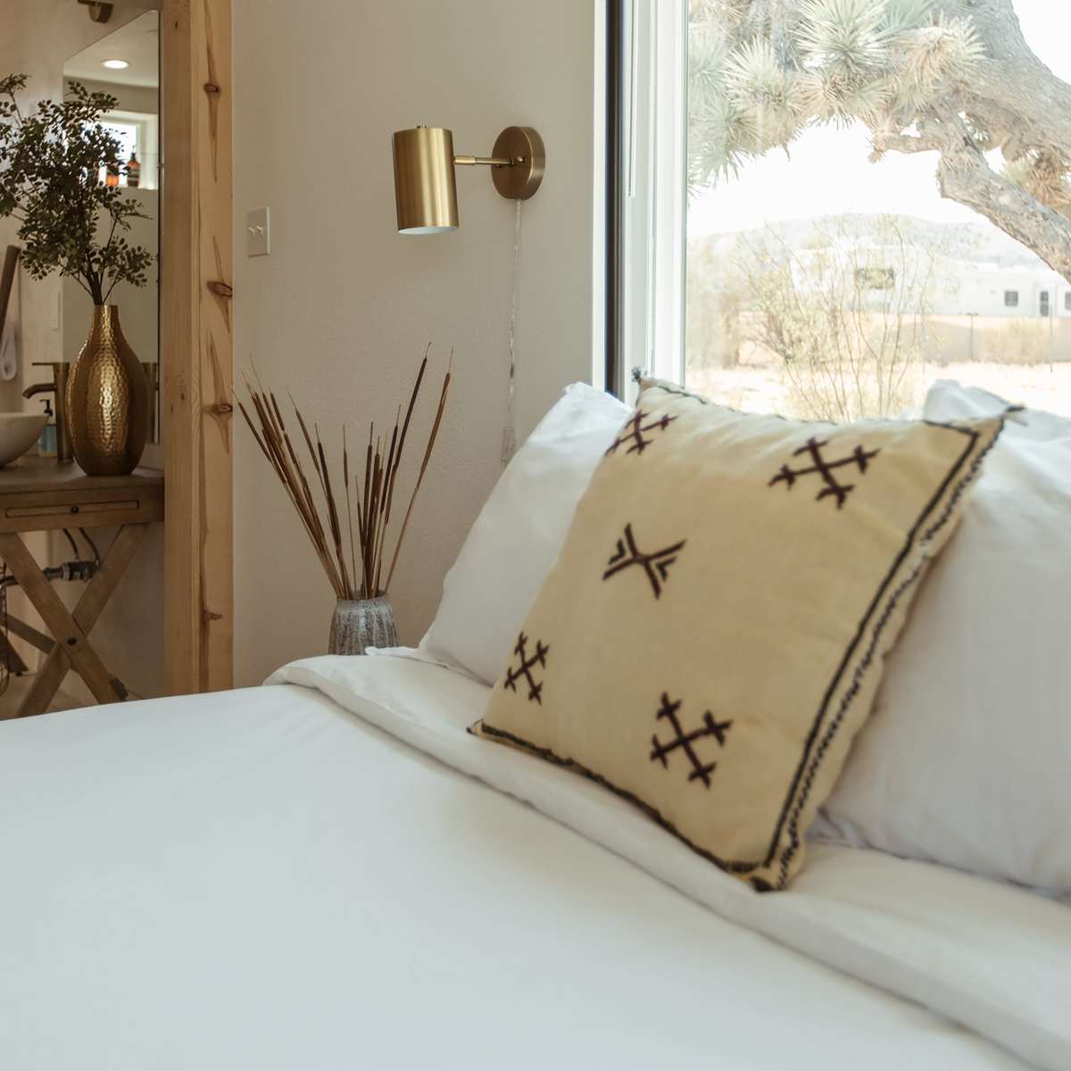 A neatly made bed with a decorative pillow, a small table with a vase and dry plants, and a window revealing a desert landscape with a Joshua tree.