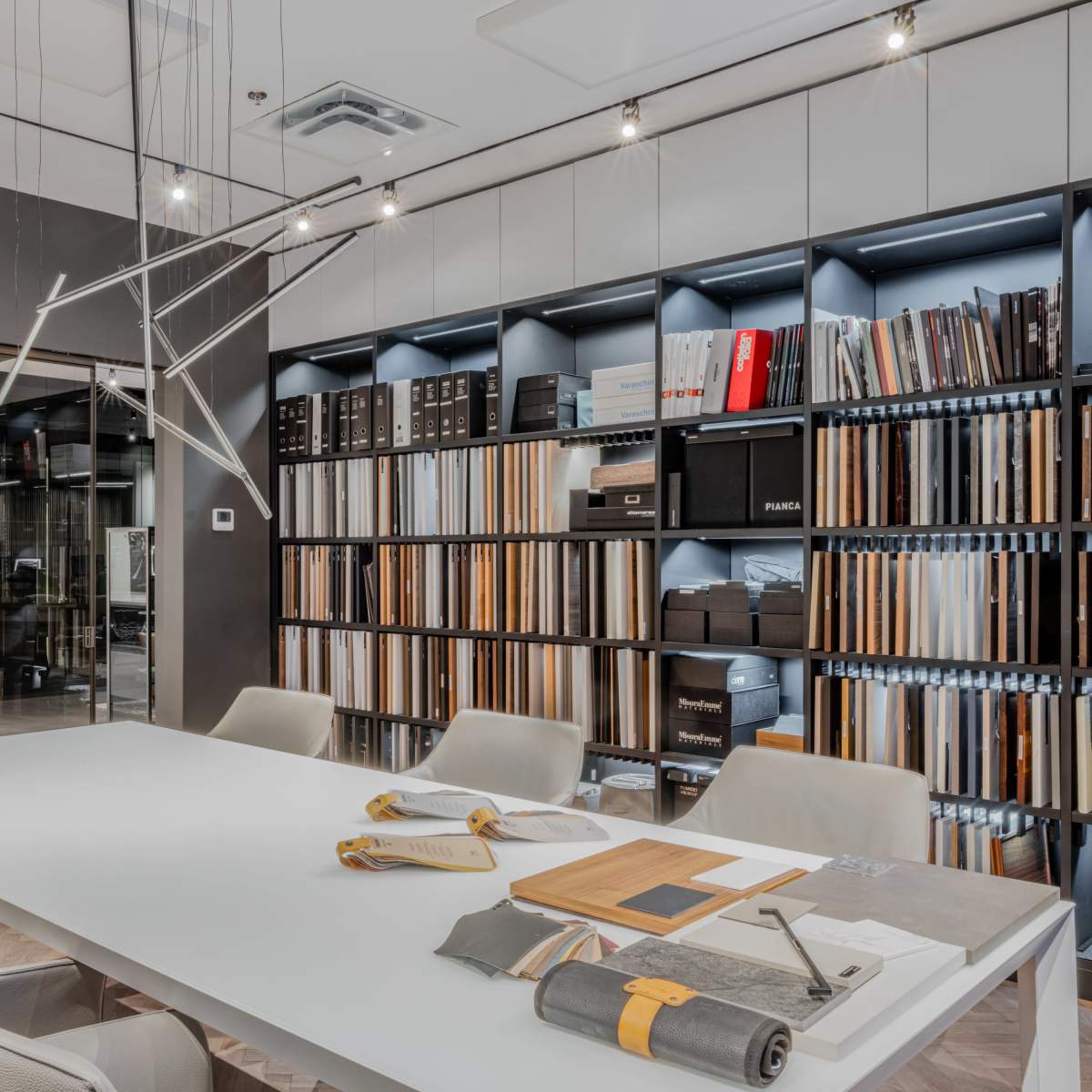 A modern office space features a large white table surrounded by light-colored chairs, with a wall of shelves displaying various binders and materials behind it.