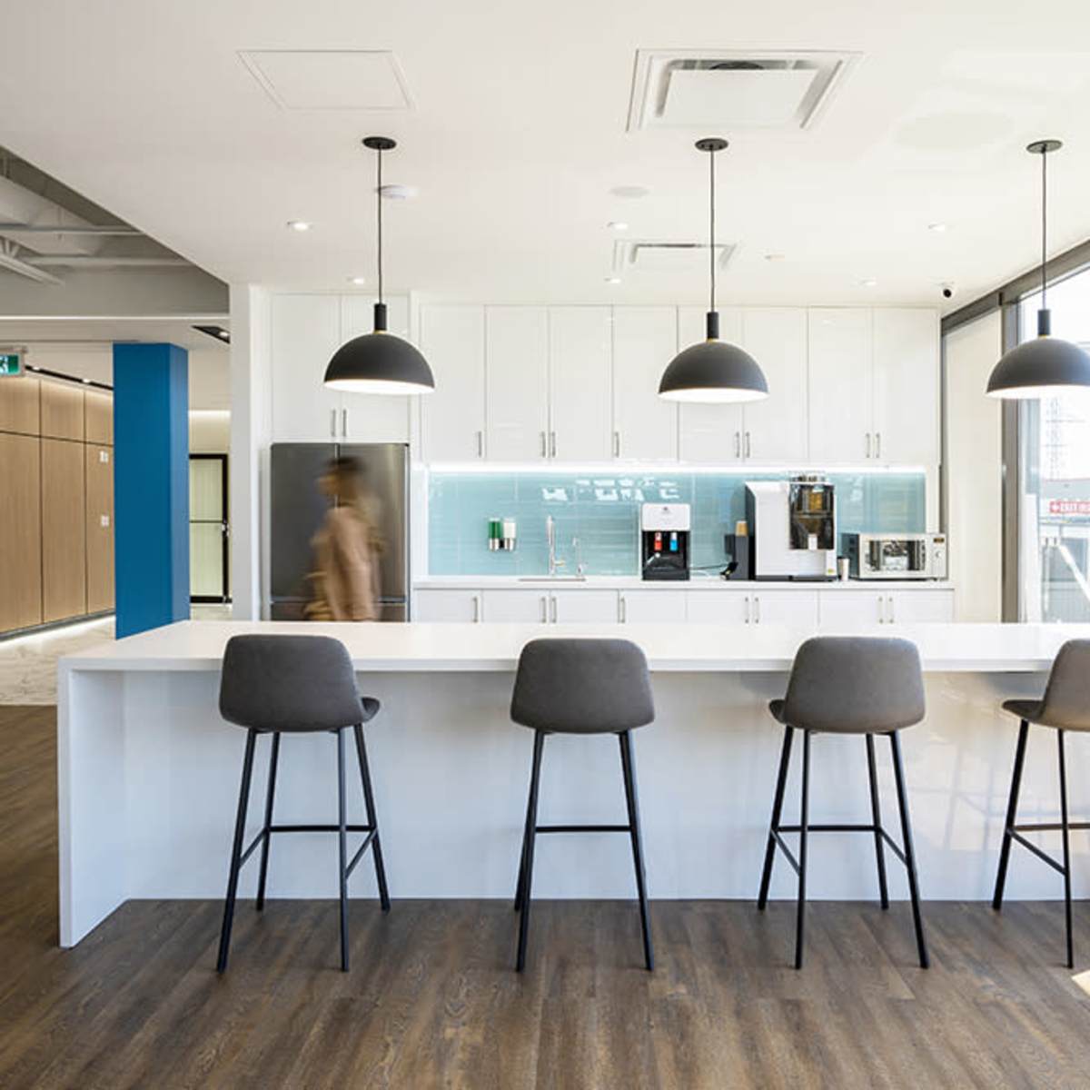 A modern kitchen with a white countertop and four bar stools, featuring a coffee machine and other appliances against a light blue backsplash.