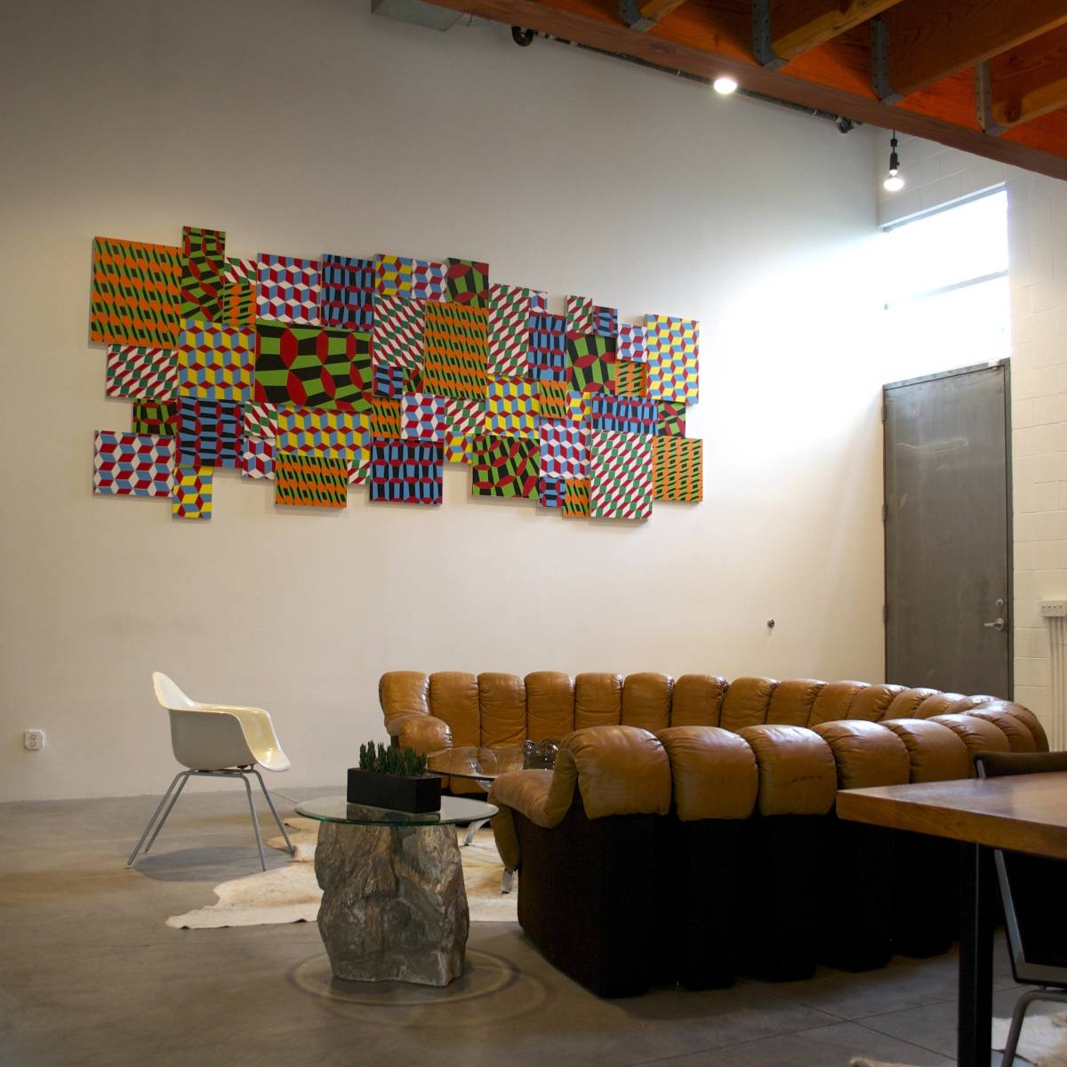 A modern living space featuring a curved brown leather sofa, a small table, and a vibrant wall art arrangement made of colorful patterned panels.