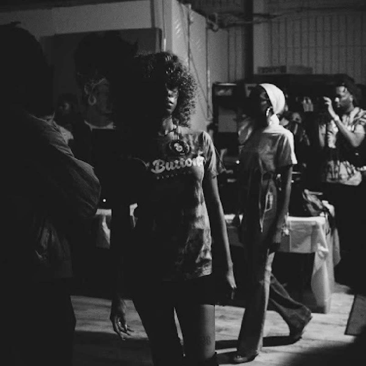A woman with curly hair walks confidently through a dimly lit room filled with people and artwork.