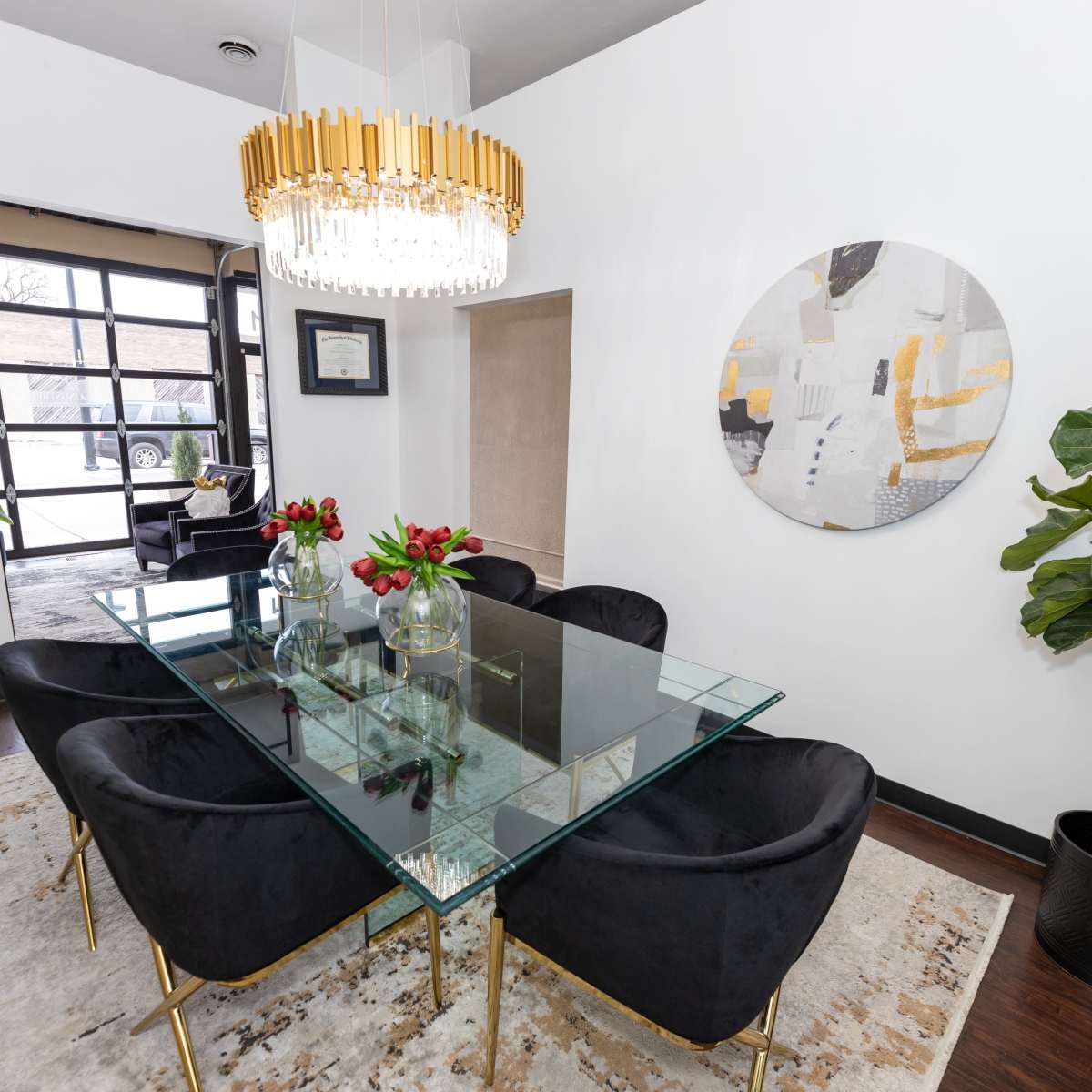 A modern dining room features a glass table with black chairs, a chandelier overhead, and a large circular wall art, accompanied by plants and decorative pieces.