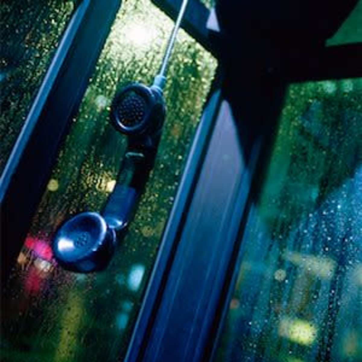 A black telephone handset hangs from a phone booth, with raindrops on the glass windows.
