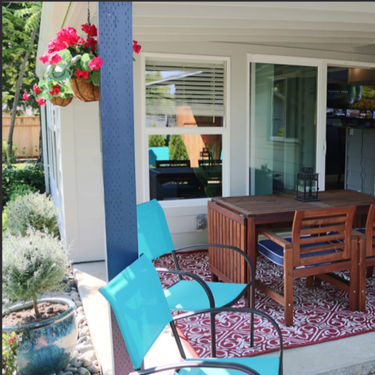 A covered patio area featuring a wooden dining table with four chairs on a patterned rug, surrounded by potted plants and a television visible indoors.