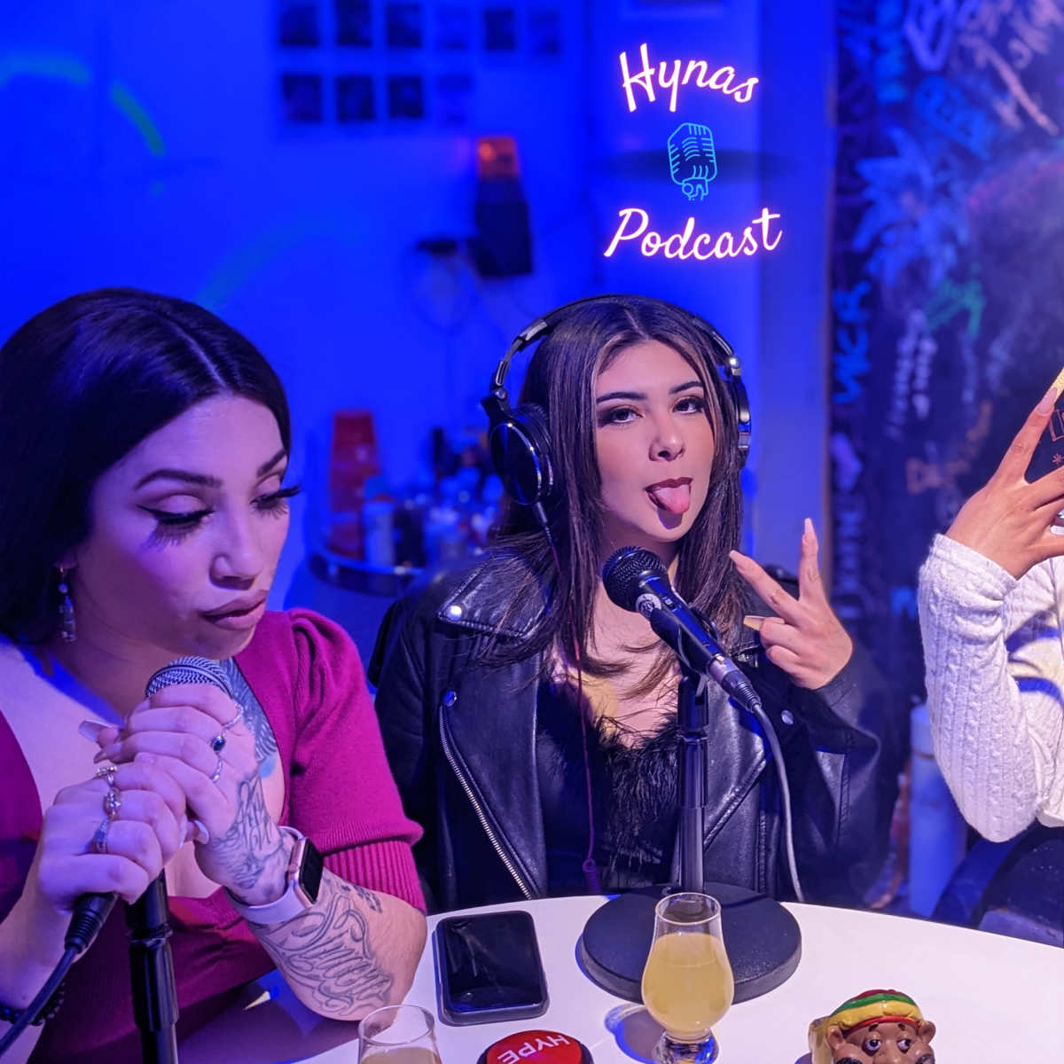 Three women sit in front of microphones in a podcast studio, with one making a playful gesture and another sticking out her tongue, while colorful lighting sets the background.