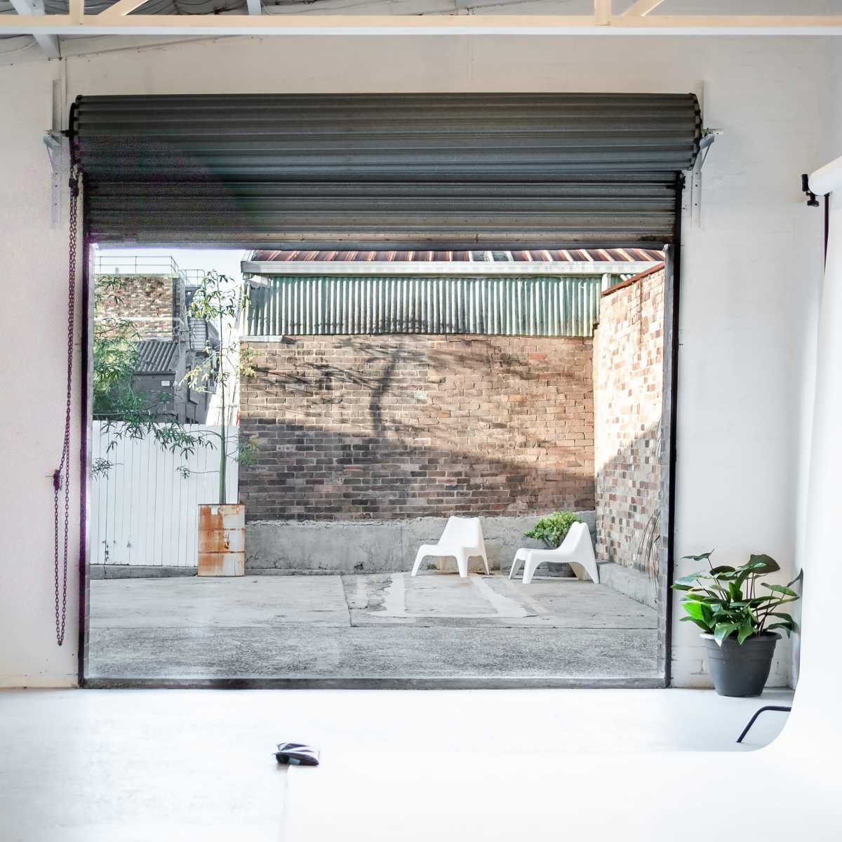 A view of a minimalist studio with an open garage door leading to a courtyard featuring a brick wall and two white chairs.