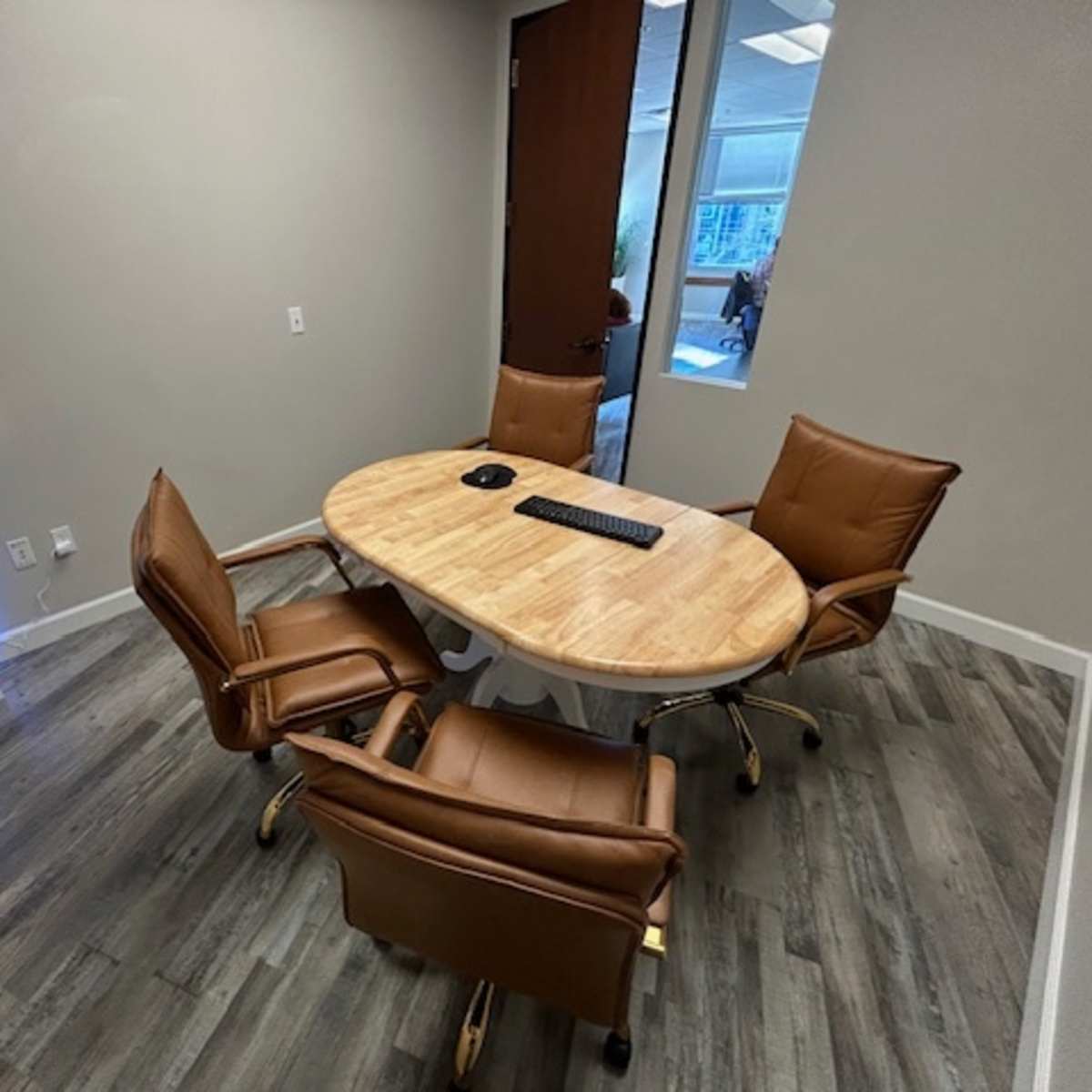 A conference room features a round wooden table surrounded by four brown leather chairs, with a keyboard on the table and a glass door in the background.