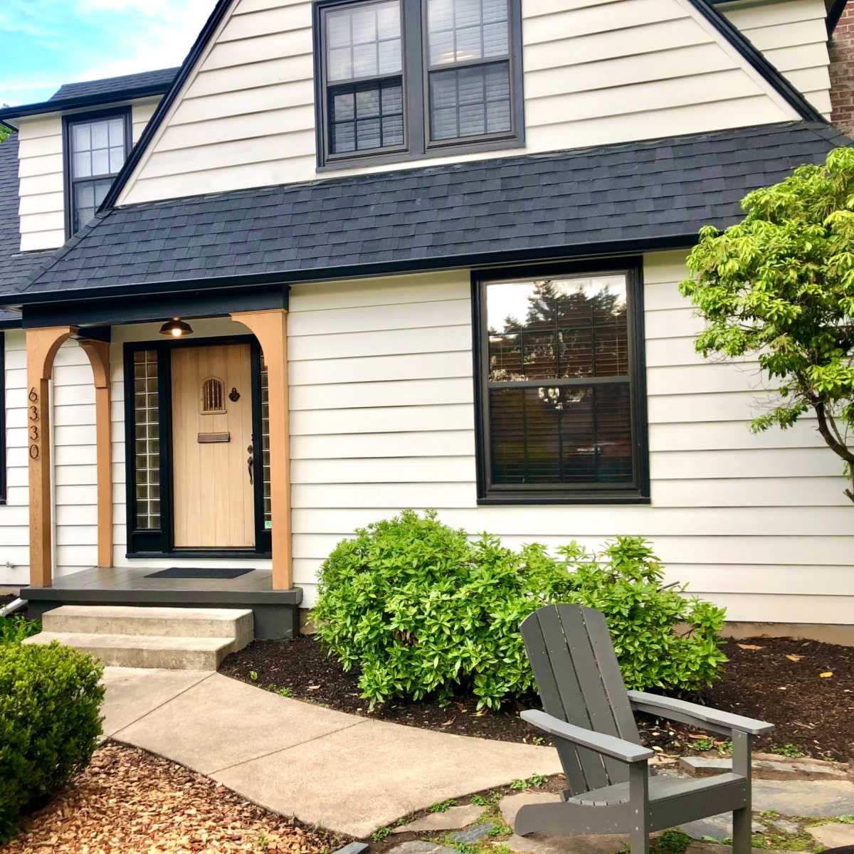 A modern, two-story house with a triangular roof, featuring a wooden front door and surrounded by well-maintained shrubs and floral beds.