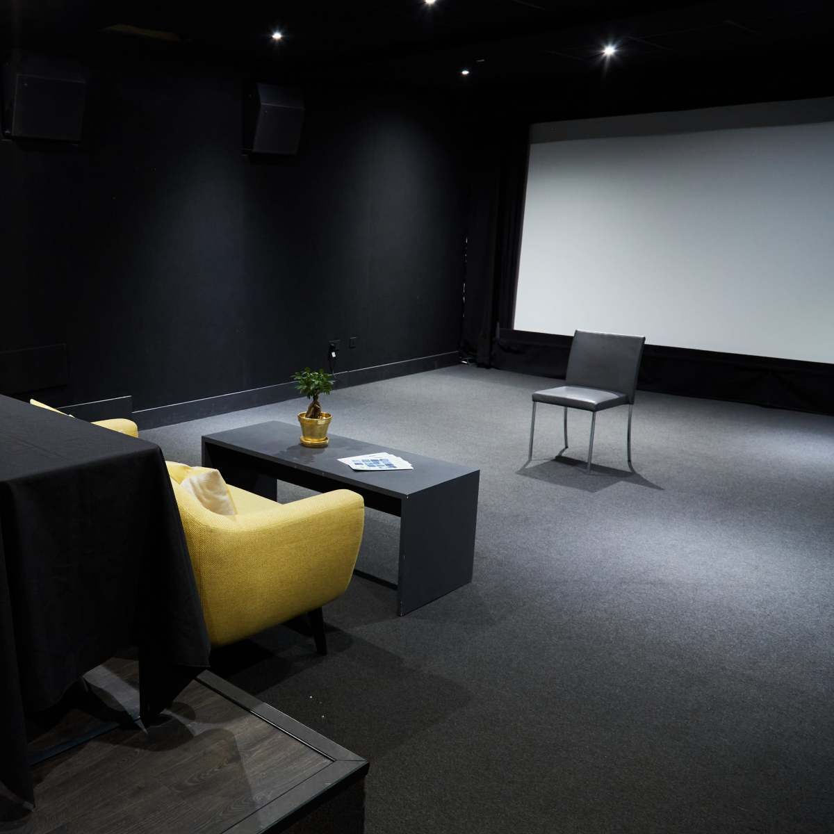 A small, dimly lit presentation space featuring a dark table, yellow armchair, black coffee table, and a single chair set against a blank white screen.