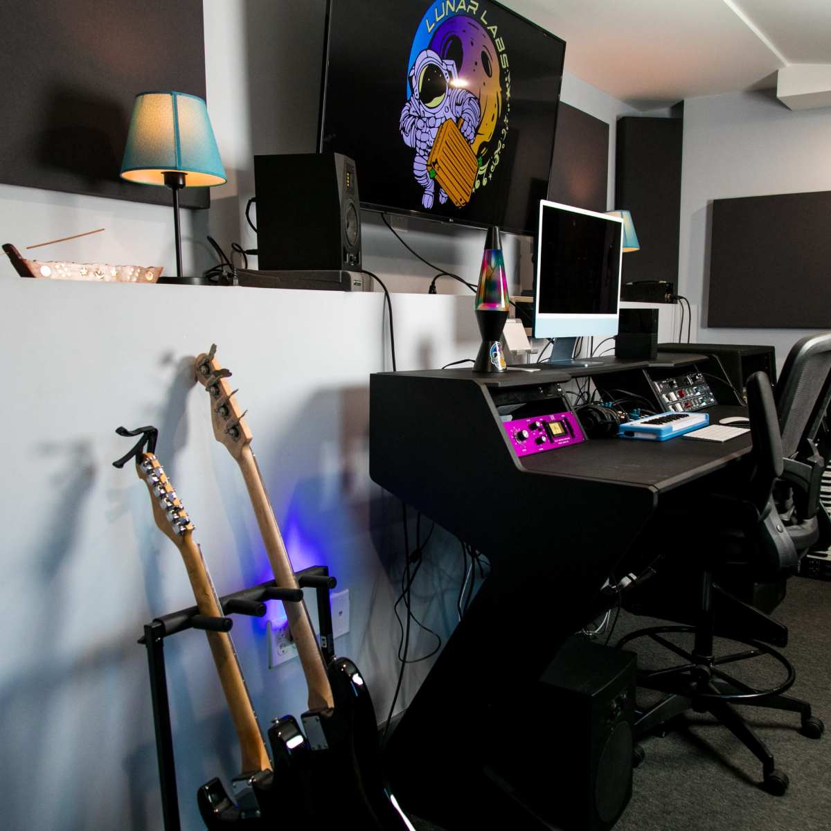 A music production studio with a large desk equipped with a computer and audio equipment, two electric guitars resting against the wall, and a microphone in the background.
