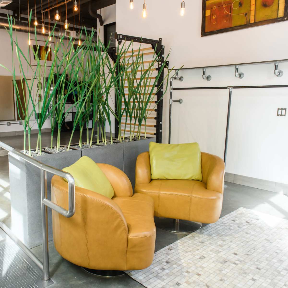 A modern lounge area featuring two curved leather chairs with green cushions, positioned near tall green plants and surrounded by large windows allowing natural light to stream in.