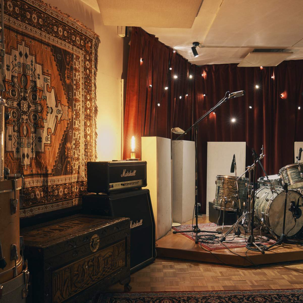 A recording studio features a drum set surrounded by soundproof panels and vintage amplifiers, with decorative rugs on the floor and walls.