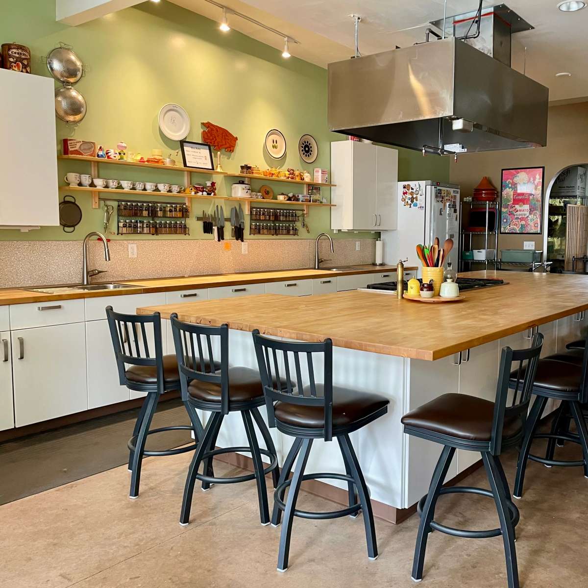 A spacious kitchen with a large wooden island surrounded by black bar stools, featuring shelves of ingredients and utensils along the walls.