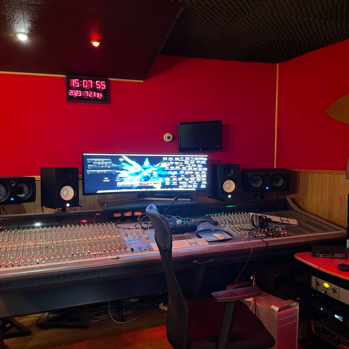 A music studio featuring a large mixing console, computer monitor, and sound equipment against a red wall and acoustic paneling.