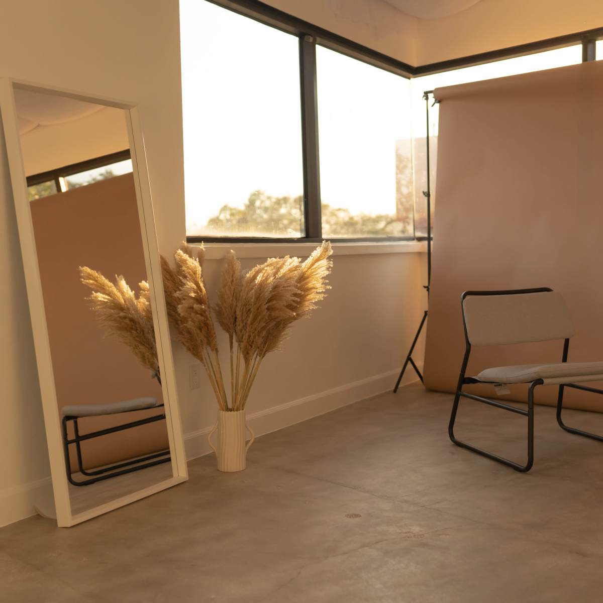 A minimalist room with a mirror, a chair, and a vase of pampas grass against a backdrop of large windows and a neutral-colored wall.