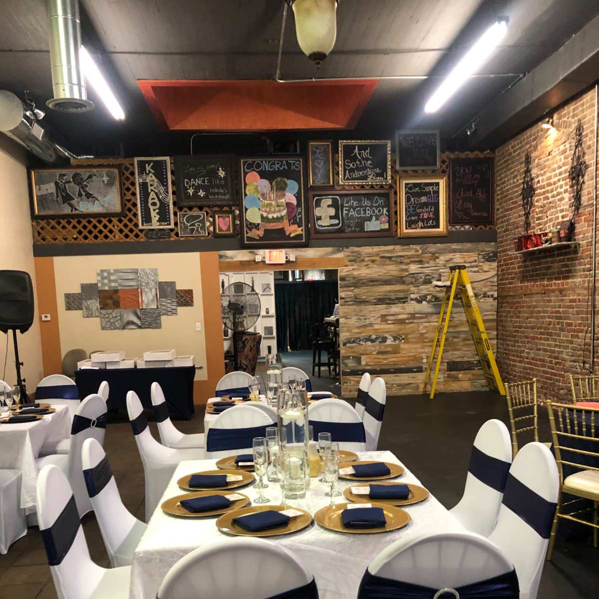 A decorated event space featuring tables set with gold chargers and dark blue napkins, surrounded by exposed brick walls and various chalkboard decorations.