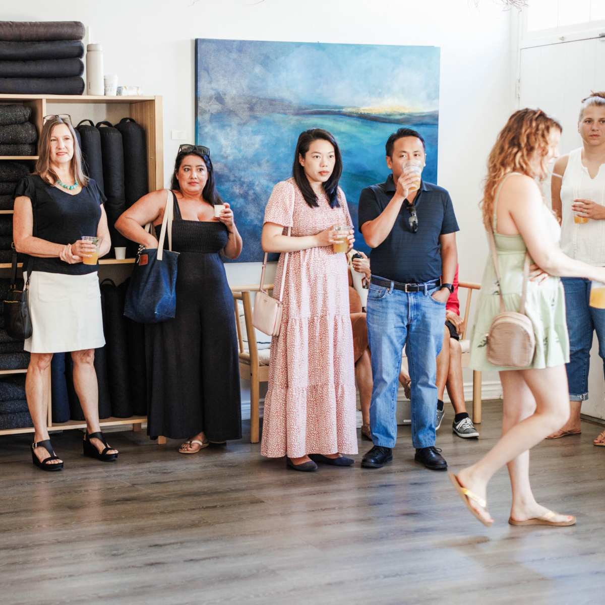 A group of people stands in a well-lit room with a shelf of folded blankets and a blue painting on the wall.