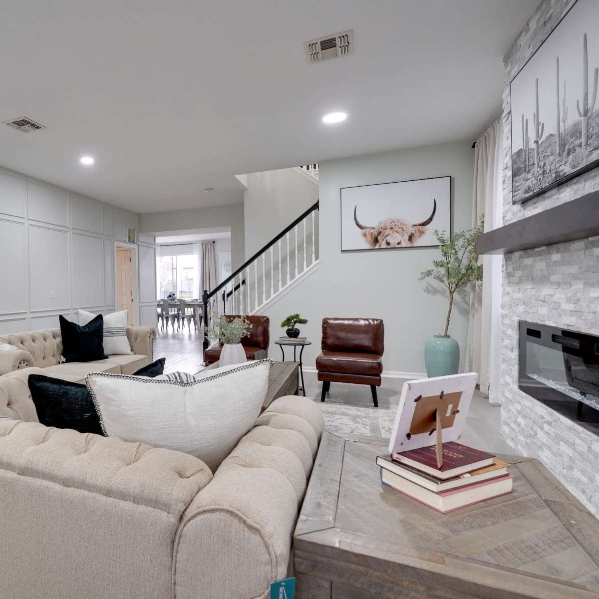 A modern living room with a light-colored tufted sofa, a coffee table with books, and a decorative plant, leading to a staircase and a dining area in the background.