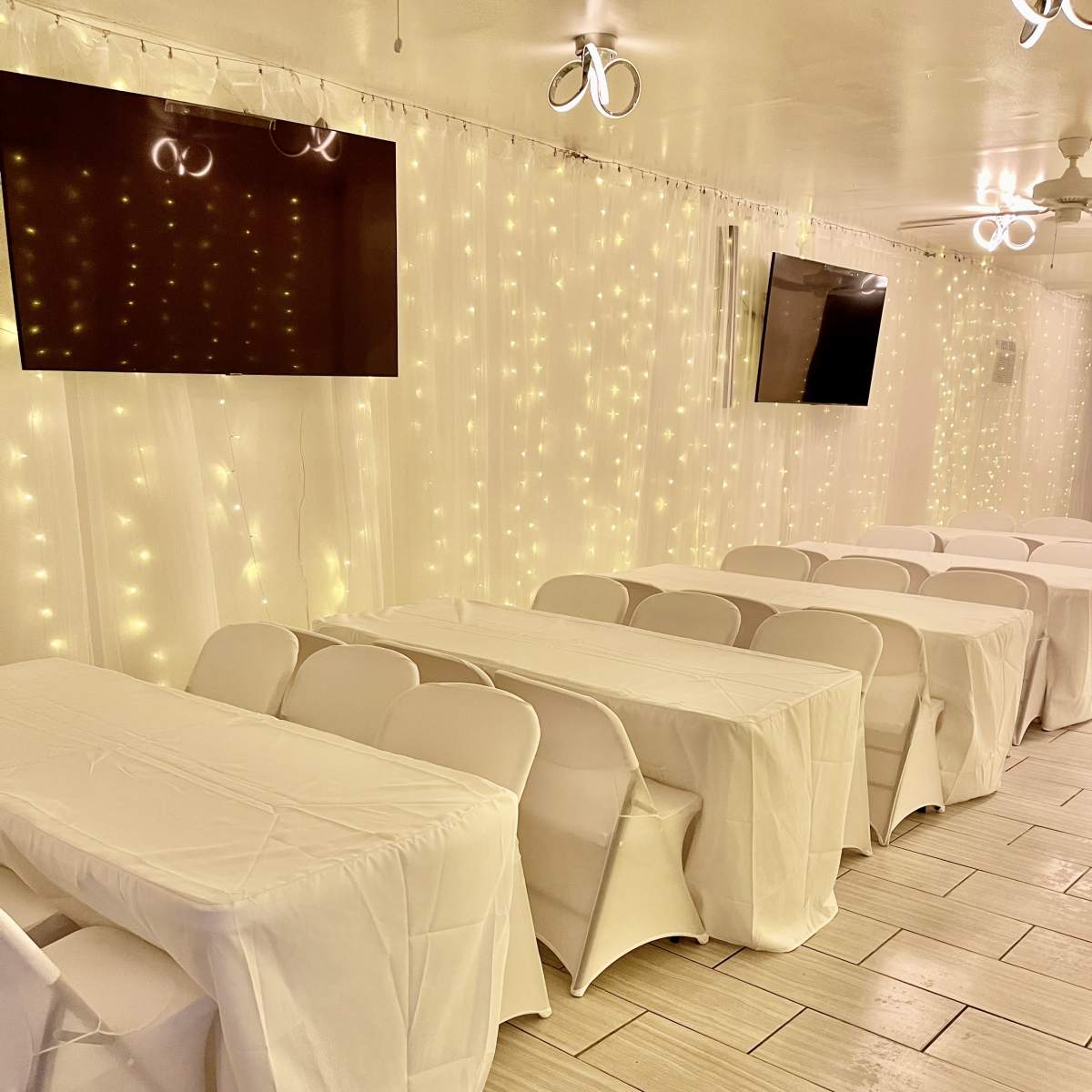 A well-lit indoor event space features neatly arranged tables covered with white cloths and wall-mounted screens in the background.
