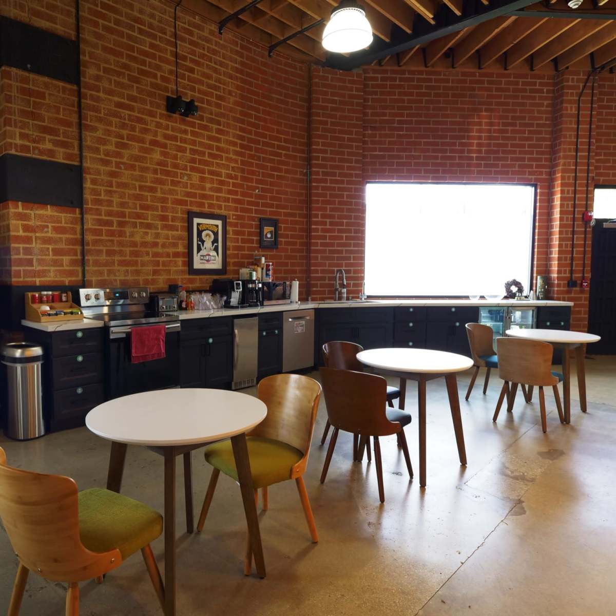 A modern kitchen space with brick walls, stainless steel appliances, and several round tables surrounded by wooden chairs.