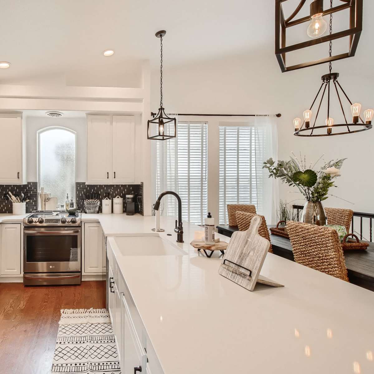 A modern kitchen with white cabinetry, a stainless steel refrigerator, a gas stove, and an island with a sink, alongside a dining area featuring woven chairs.