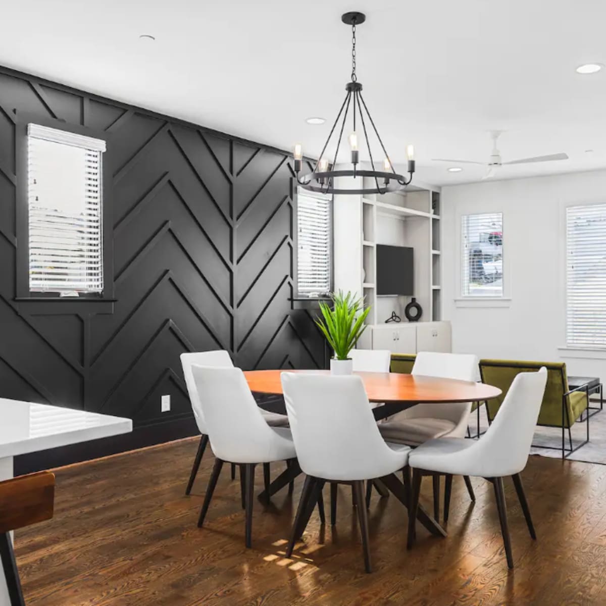 A modern dining area features a round wooden table surrounded by white chairs, with a black accent wall and large windows allowing natural light.