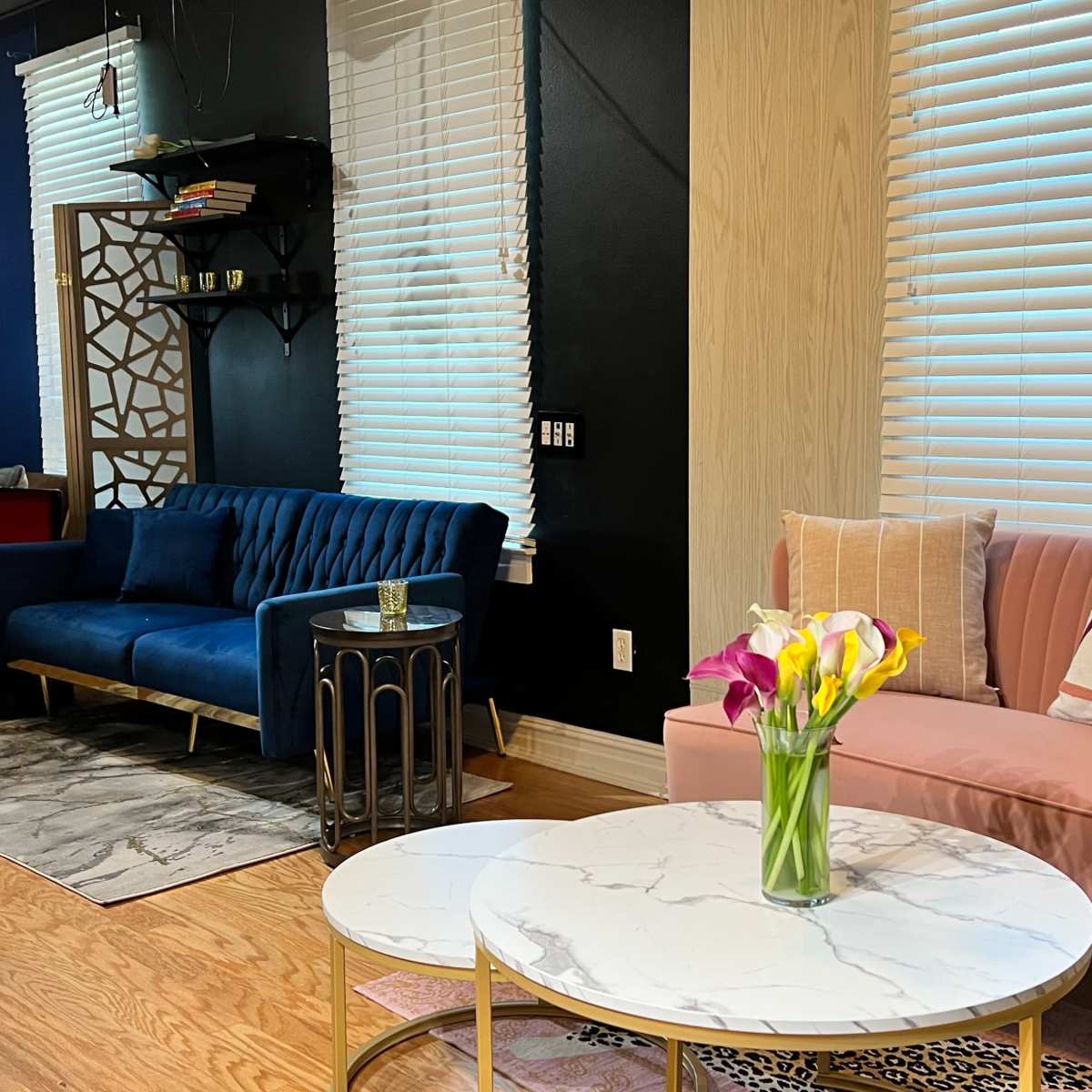 A modern living room with a blue velvet sofa, a pink armchair, and a round marble coffee table, accented by a vase of fresh flowers.