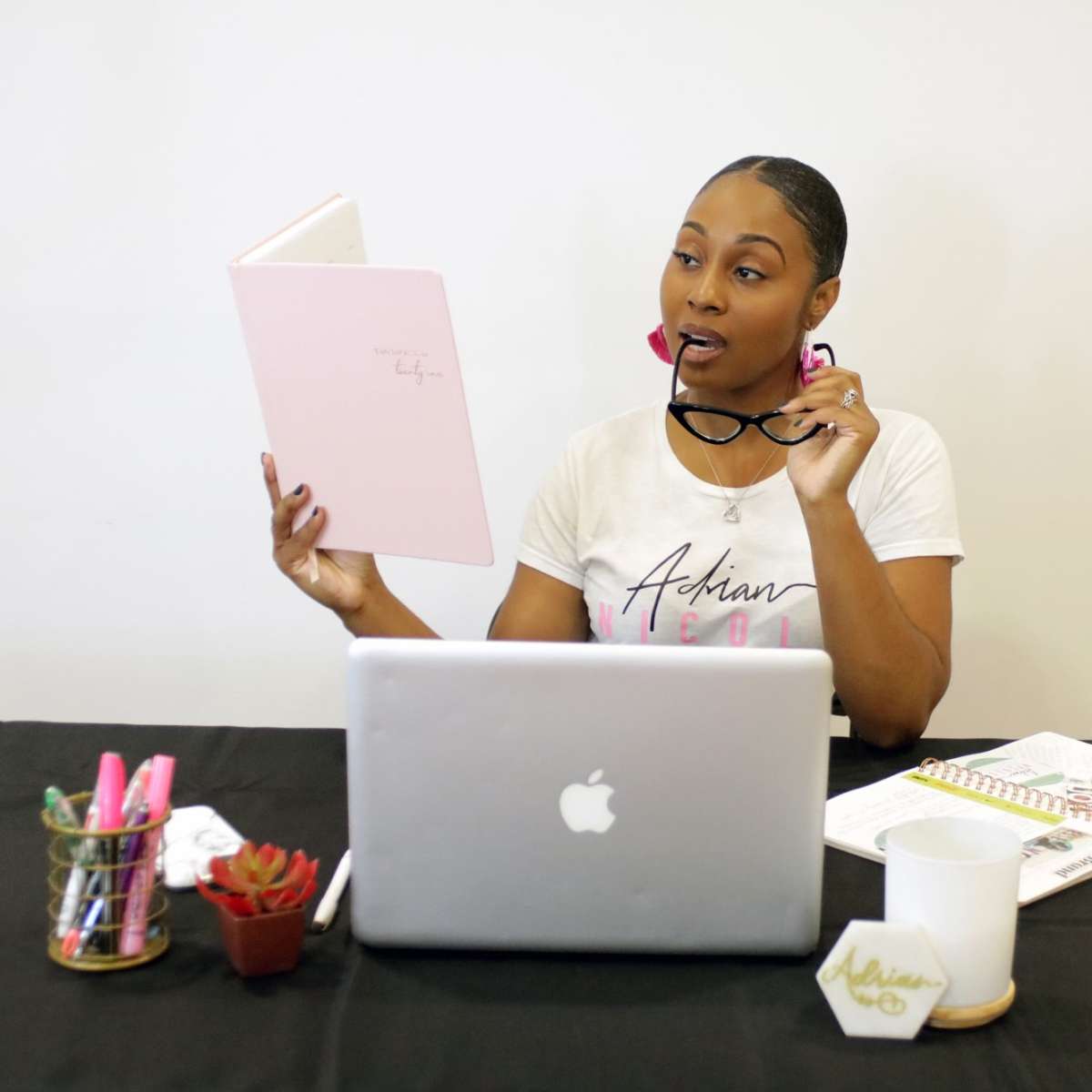 A woman is seated at a desk with a laptop, holding glasses in one hand and reviewing a pink notebook.