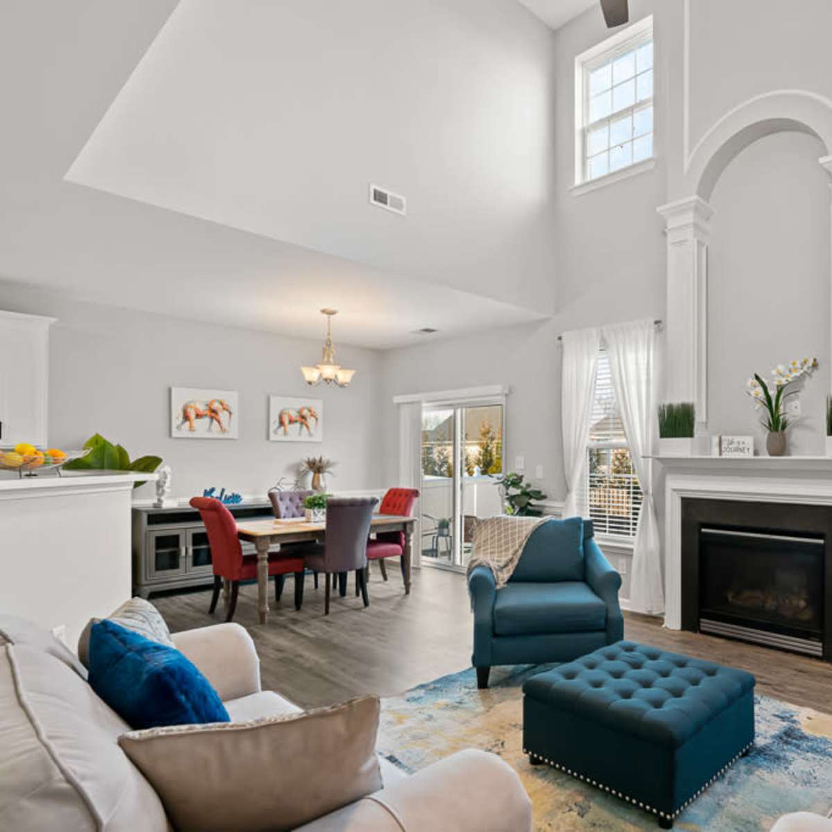 The living room features a vaulted ceiling, a fireplace with decorative molding, a blue tufted ottoman, and large windows allowing natural light.