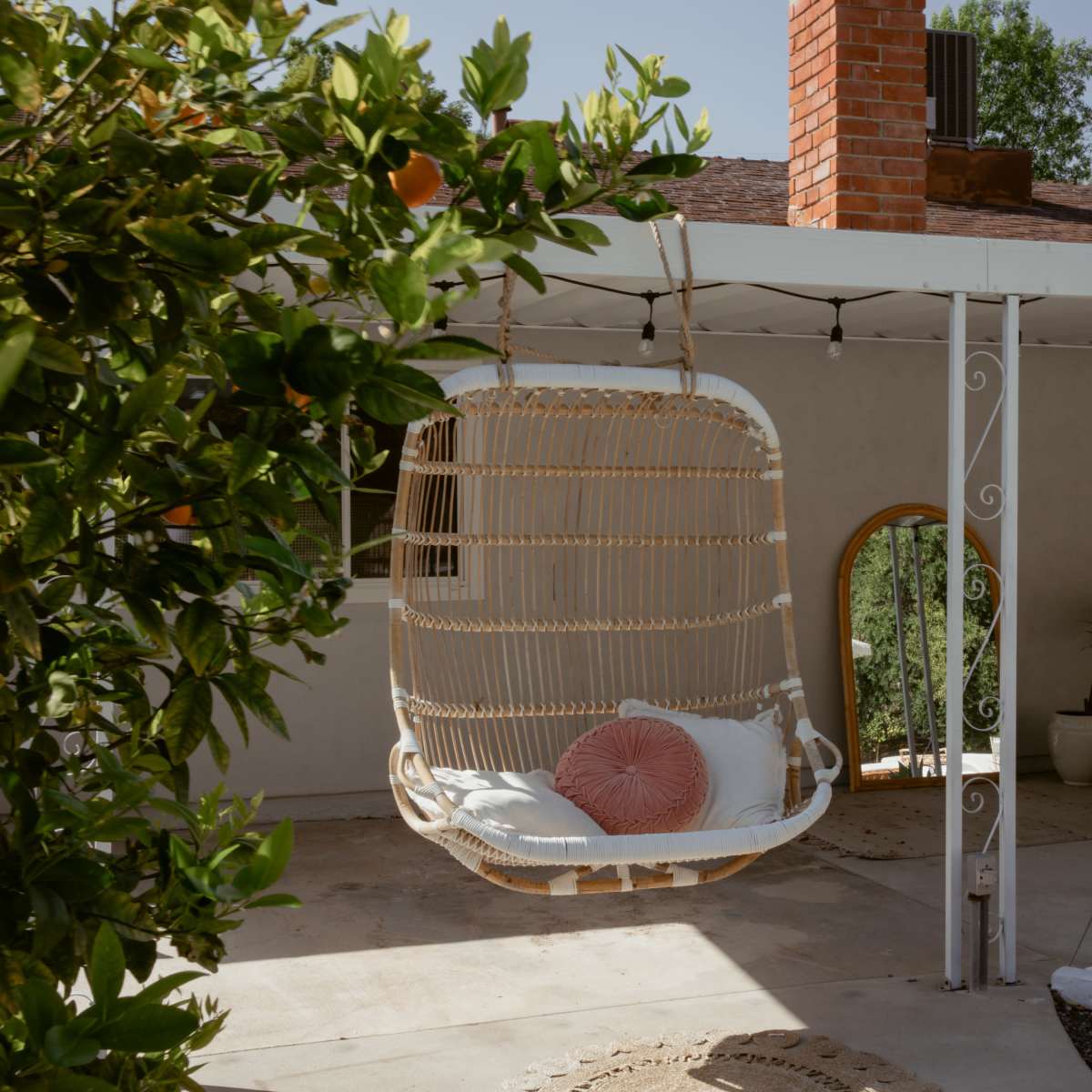 A hanging chair with a pink cushion is suspended from a covered patio, surrounded by potted plants and a view of a house with a brick chimney.