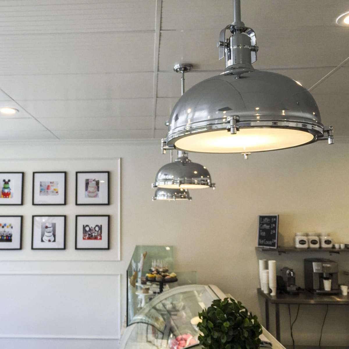 A brightly lit café interior with chrome pendant lights and framed artwork on the wall, alongside a display of pastries.