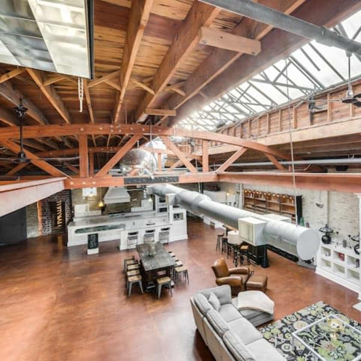 A spacious, loft-style living area with exposed wooden beams, large windows, and an open kitchen adjacent to a dining table and seating area.