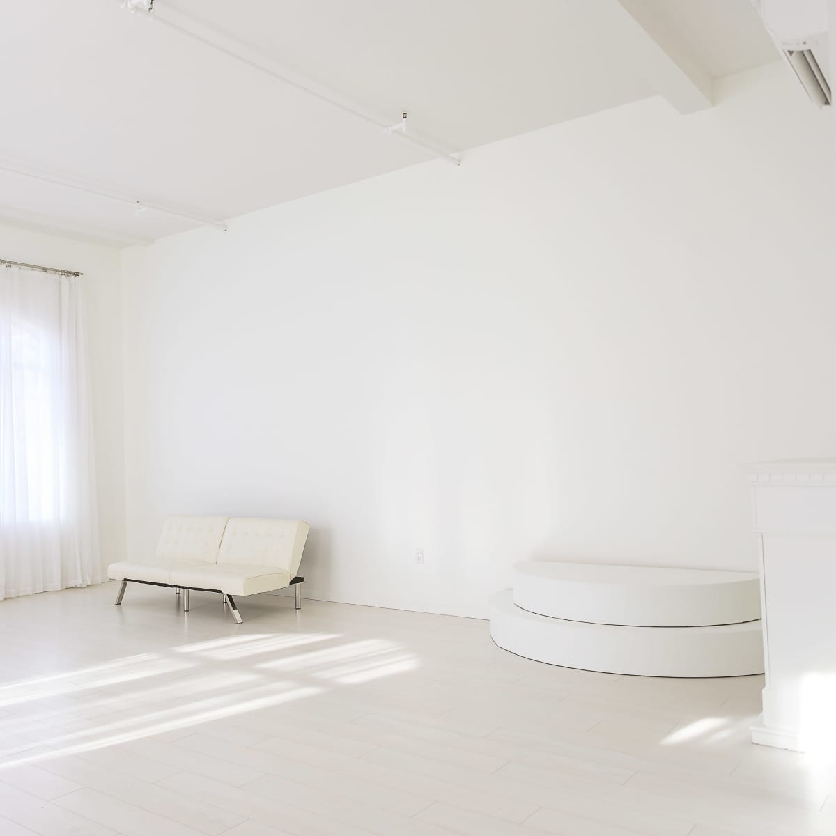 A bright, empty room features a white sofa, a curved white platform, and large windows covered with sheer curtains.