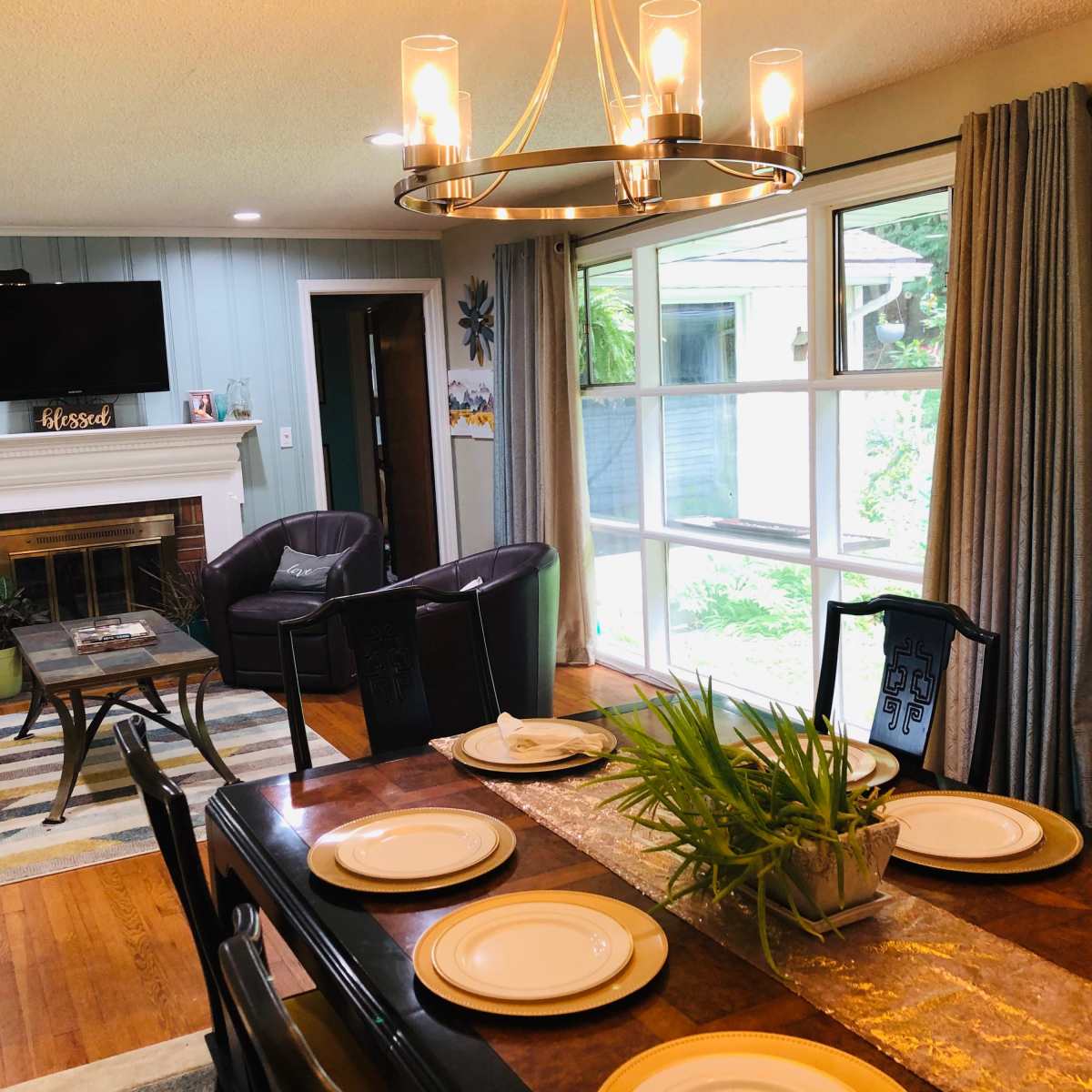 A dining area with a wooden table set for a meal, surrounded by chairs, adjacent to a living space featuring a fireplace and a large window overlooking a garden.