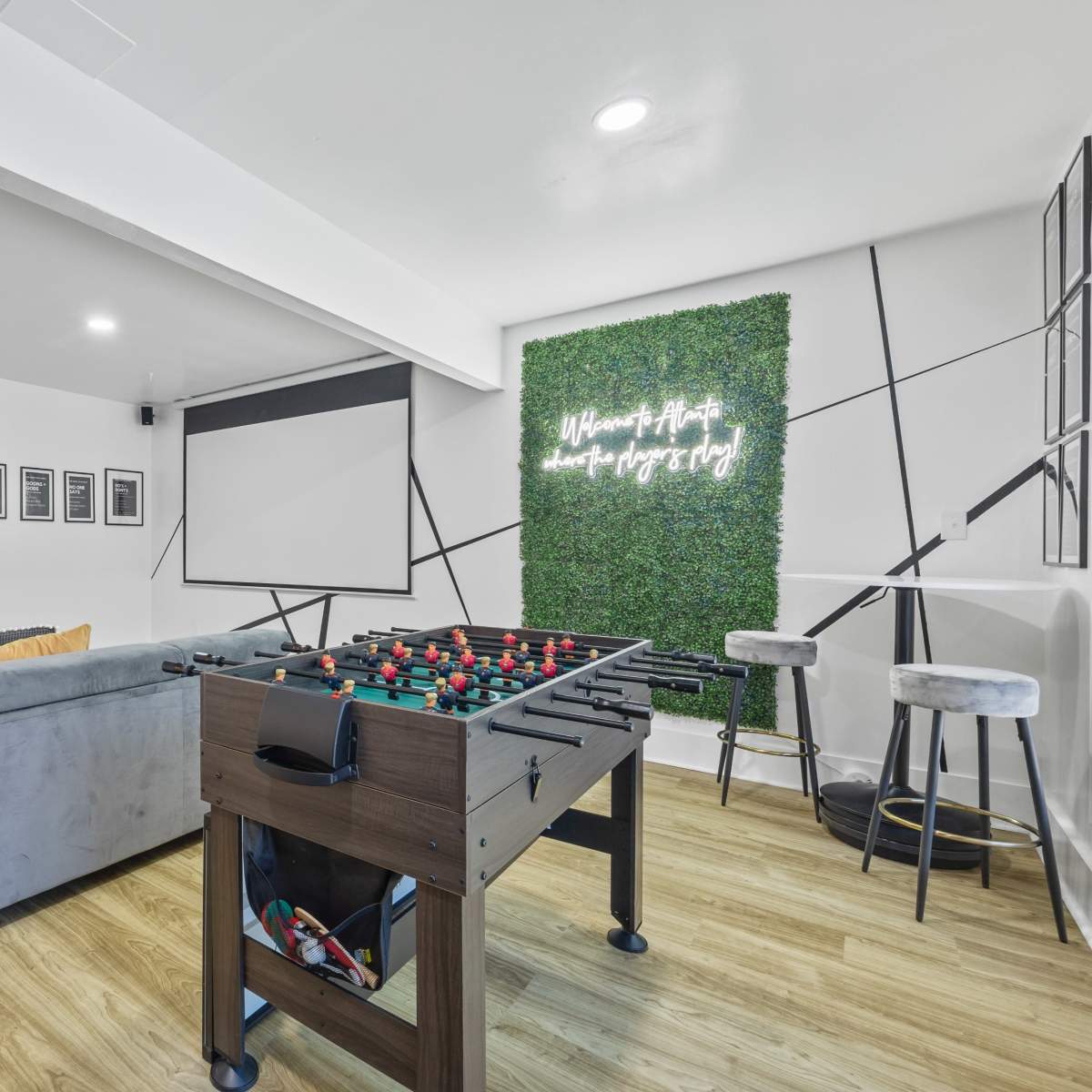 A modern game room featuring a foosball table, a gray sectional sofa, a projector screen, and a green accent wall with decorative text.