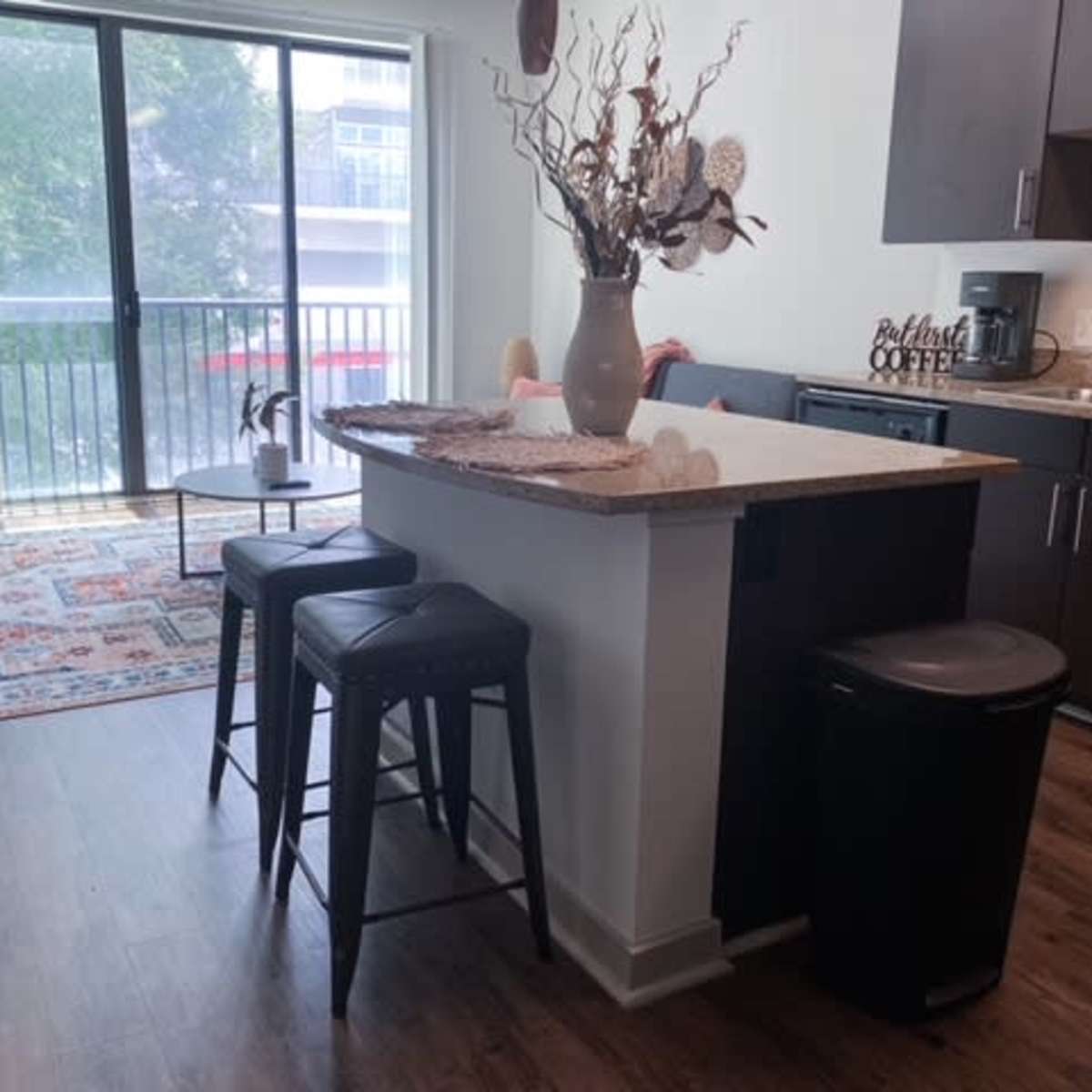 A kitchen area features a bar with three stools, a decorative vase on the countertop, and a sliding glass door leading to a balcony.