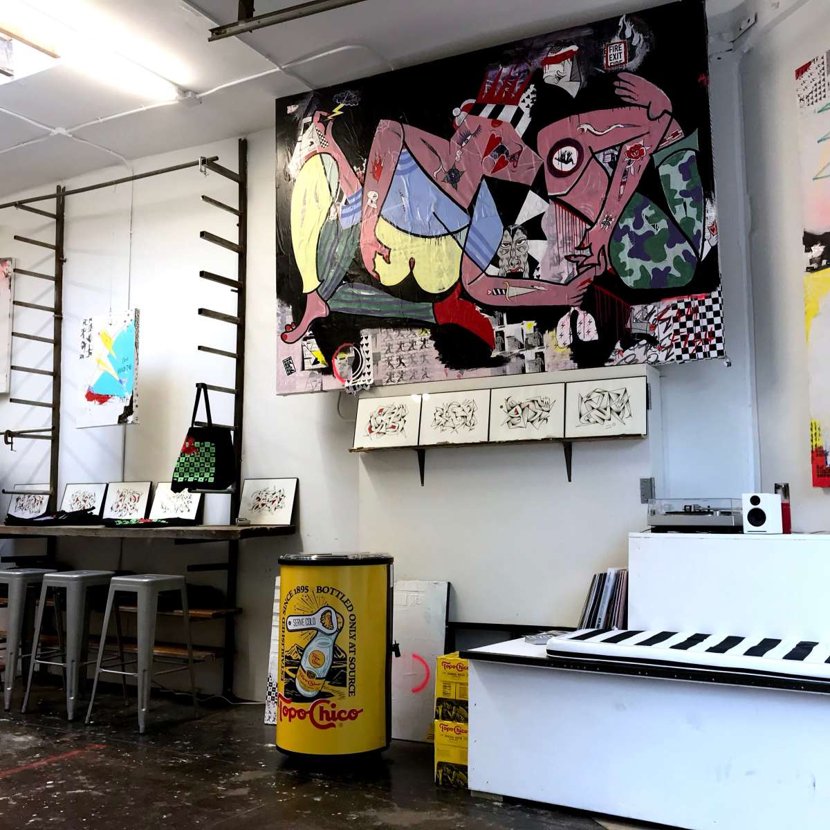 An art studio with colorful paintings on the walls, various framed artworks displayed on shelves, and merchandise such as T-shirts and a yellow trash can in the foreground.