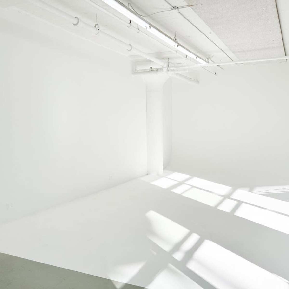 A spacious, empty room with white walls and a seamless white floor, illuminated by natural light streaming through large windows.