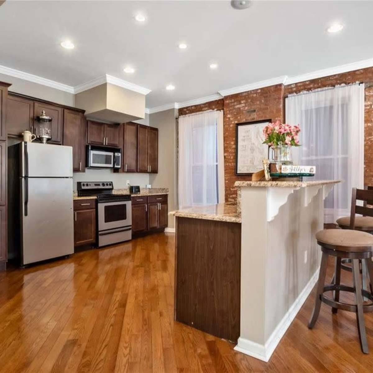 The kitchen features a mix of stainless steel appliances, dark wooden cabinetry, and a brick accent wall, with a breakfast bar and two stools.