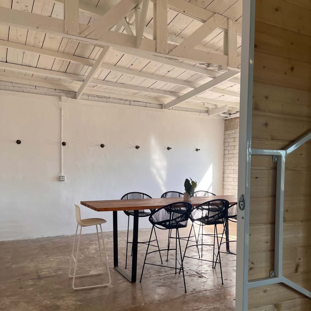 A minimalistic meeting room features a wooden table surrounded by black wire chairs, with natural light illuminating the space through exposed beams and a white wall.