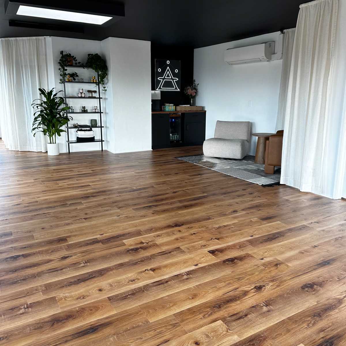 A spacious interior room with wooden flooring, white curtains, a dark wall featuring a geometric design, and a small bookshelf against one side.
