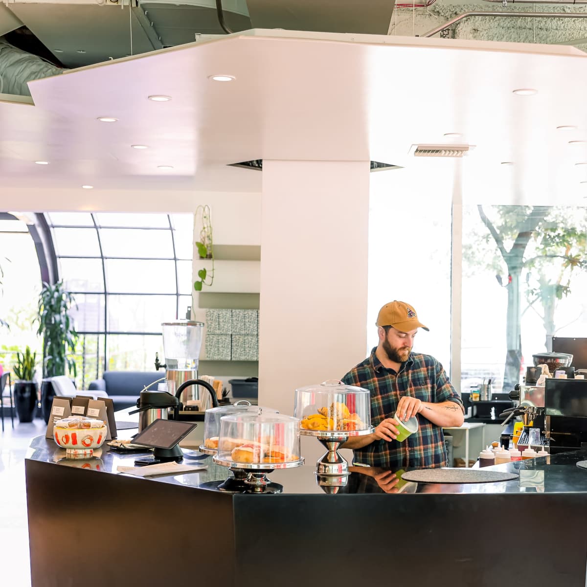 A barista wearing a cap prepares drinks at a modern café counter with glass display cases.